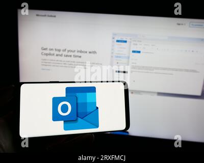 Persona che possiede il cellulare con il logo del software di posta elettronica Microsoft Outlook sullo schermo davanti alla pagina Web dell'azienda. Concentrarsi sul display del telefono. Foto Stock