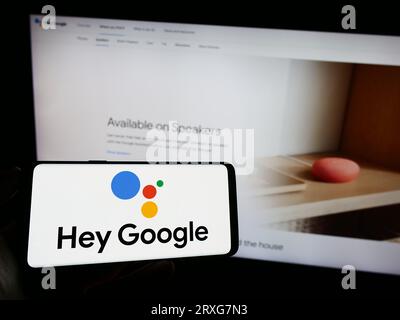 Persona che tiene il cellulare con il logo del software di assistenza virtuale Google Assistant sullo schermo davanti alla pagina Web. Concentrarsi sul display del telefono. Foto Stock