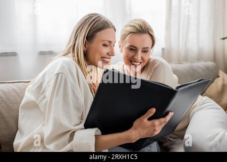 belle sorelle in un abbigliamento informale sedute su un divano che guardano un album fotografico, un legame con la famiglia Foto Stock
