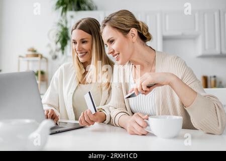 due bellissime sorelle bionde in un abbigliamento alla moda con carta di credito e una tazza di tè che guardano il computer portatile Foto Stock