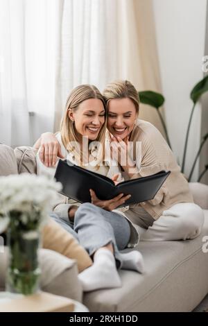 belle e belle sorelle in un abbigliamento alla moda che ridono e guardano album fotografici, legami familiari Foto Stock