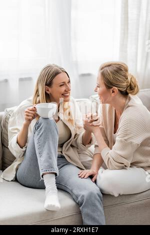 due adorabili sorelle in abiti pastello alla moda sedute sul divano bevendo il tè tenendosi per mano, legando Foto Stock