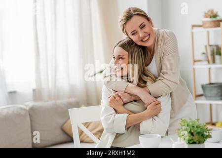 bellissime sorelle in un abbigliamento alla moda che abbracciano e sorridono sinceramente sullo sfondo della cucina, legando Foto Stock