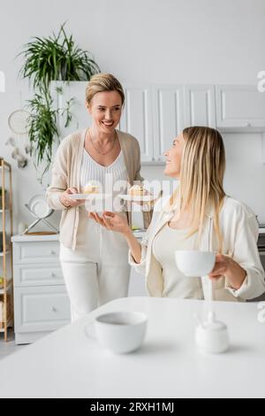 due sorelle in un bell'abbigliamento pastello che parlavano e sorridono, cupcake in mano, legame con la famiglia Foto Stock