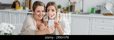 due sorelle bionde in un bell'abbigliamento che fanno selfie sullo sfondo della cucina, legano la famiglia, striscione Foto Stock