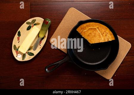 Piatto di padella in ghisa nera con pane di mais appoggiato su una piastra calda con teglia di burro e coltello su un lato Foto Stock