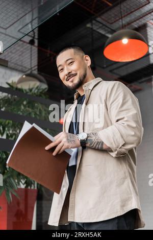 simpatico lavoratore asiatico con tatuaggio che tiene la cartella e guarda la macchina fotografica vicino alla porta di vetro Foto Stock