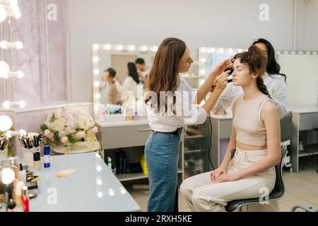 Ampia foto di una parrucchiera con pinze per capelli che arricciano i capelli di una giovane donna in uno studio di bellezza. Truccatore che applica fondamenti su faccia femminile usando Foto Stock