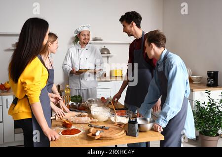 Italian chef with prepared pizza and group of young people after cooking class in kitchen Stock Photo