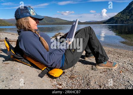 Donna che vive la sua vita migliore leggendo un libro sulle rive di un remoto lago Yukon; Yukon, Canada Foto Stock