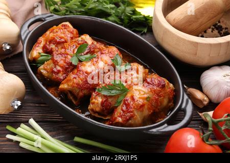 Deliziosi involtini di cavolo ripieni cucinati con salsa di pomodoro e ingredienti su un tavolo di legno, primo piano Foto Stock