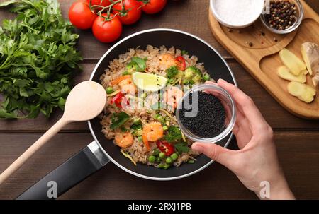 Donna che aggiunge semi di sesamo al riso con gamberetti e verdure al tavolo di legno, vista dall'alto Foto Stock