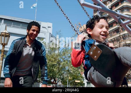 Padre single afro latino venezuelano che gioca con suo figlio a swing nel parco pubblico, sorridendo felice, spazio copia, concetto di stili di vita. Foto Stock