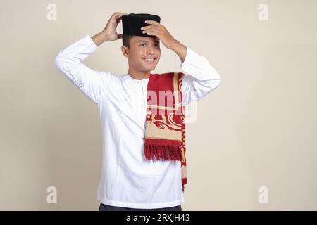 Ritratto di un attraente uomo musulmano asiatico in camicia bianca con berretto che cerca di regolare il suo songkok o skullcap nero. Immagine isolata su sfondo grigio Foto Stock