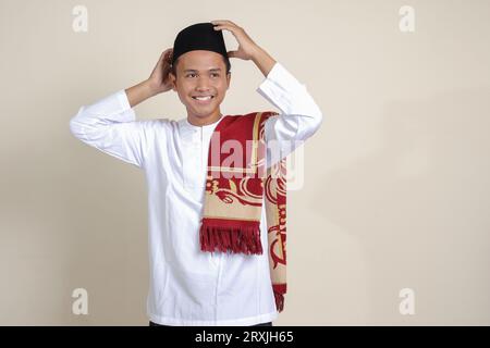 Ritratto di un attraente uomo musulmano asiatico in camicia bianca con berretto che cerca di regolare il suo songkok o skullcap nero. Immagine isolata su sfondo grigio Foto Stock