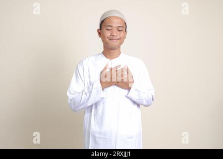 Ritratto di un attraente uomo musulmano asiatico in camicia bianca con berretto che mette la mano sul cuore, sentendosi molto grato. Immagine isolata su sfondo grigio Foto Stock