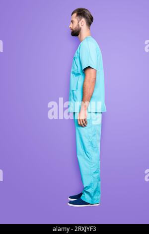 Bottoncino a figura intera, mezza faccia vista laterale ritratto di bell'uomo attraente con setole, acconciatura in tuta da laboratorio blu, isolato su sfondo grigio Foto Stock