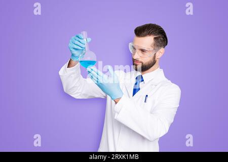 Ritratto di scienziato occupato concentrato con stoppia in bianco camice da laboratorio, guanti esaminare, guardando il pallone con liquido multicolore nel braccio Foto Stock