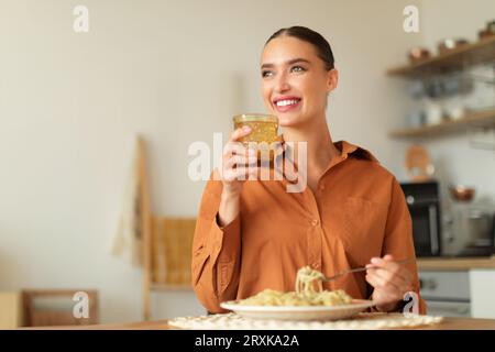 Una donna felice che tiene un bicchiere d'acqua, che mangia spaghetti fatti in casa, si siede al tavolo in cucina e guarda da parte, spazio libero Foto Stock