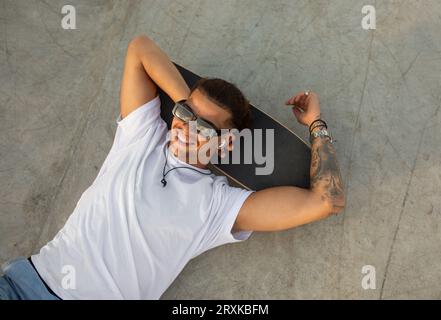 Elegante uomo tatuato che si rilassa sullo skateboard e ascolta la musica in padiglioni, si rilassa nello skate Park, vista dall'alto Foto Stock