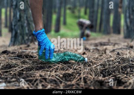 Il primo piano cattura una mano con i guanti che raccoglie un flacone di plastica scartato. Sullo sfondo, sotto il baldacchino degli alberi, un altro individuo si piega verso ga Foto Stock