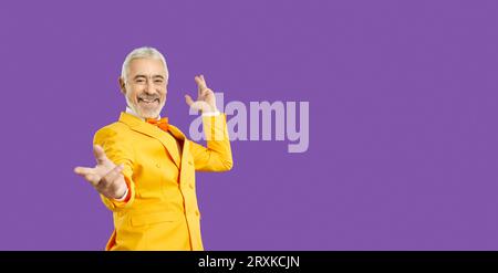 Anziano con i capelli grigi, l'uomo allegro sorriso estende la mano alla fotocamera indossa un vestito giallo e sta in studio Foto Stock