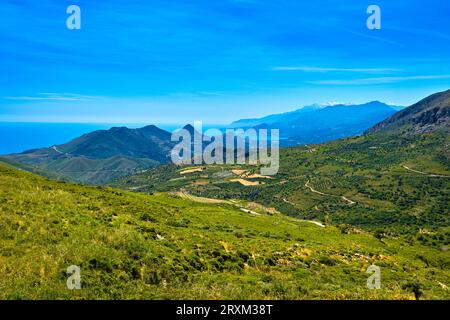 Paesaggio estivo dell'isola di Creta, Grecia. Alte montagne, cime innevate, verdi colline, strade e piantagioni di ulivi, erba alta, vegetazione e vegetazione lussureggianti. Mare Mediterraneo, cielo blu e nuvole. Panorama idilliaco nelle giornate di sole. Foto Stock