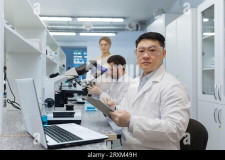 Un gruppo internazionale di giovani scienziati lavora in laboratorio. Un uomo asiatico è seduto a una scrivania e sta usando un tablet, dietro il quale i colleghi stanno esaminando con un microscopio. Foto Stock