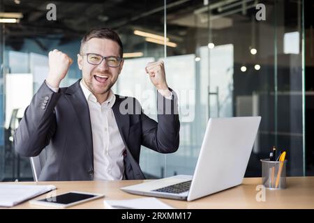 Ritratto di un giovane uomo d'affari felice seduto a una scrivania in ufficio e che celebra il successo sul lavoro. Foto Stock