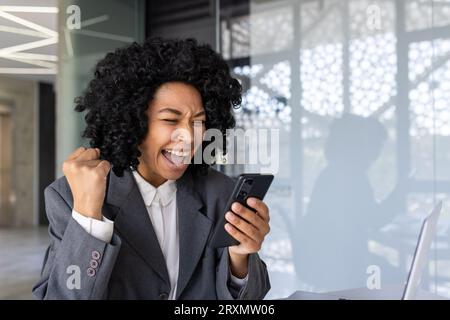 La donna afro-americana in primo piano all'interno dell'ufficio sul posto di lavoro ha ricevuto una notifica Internet online WIN Message, una donna d'affari che utilizza un'app sullo smartphone per celebrare il trionfo e ottenere risultati. Foto Stock