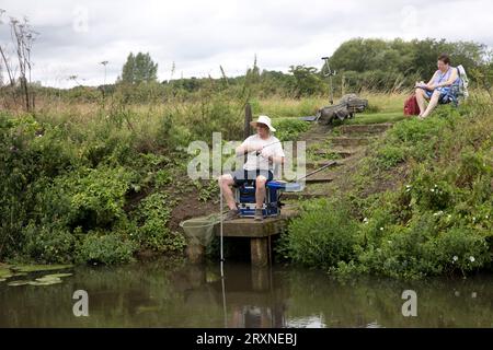 Uomo seduto su una piattaforma rialzata che pesca con canna e lenza sul fiume Avonwith, moglie seduta pazientemente readibita vicino a Evesham, Regno Unito Foto Stock