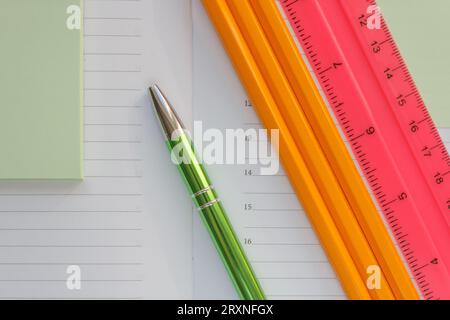 Primo piano di un diario aperto per scrivere, una penna verde, tre matite arancioni e un righello rosa con una scala. cancelleria sul tavolo Foto Stock