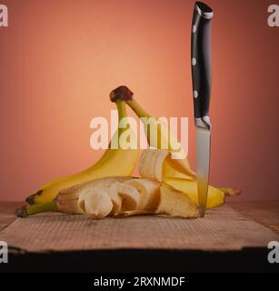 Mazzo di banane con una che è stata aperta e tagliata su una superficie di legno con un coltello sul lato. Foto Stock