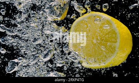 Sott'acqua di limonata dolcificata appena spremuta che affetta di limoni crudi cadendo in acqua soda su sfondo blu scuro o nero. Primo piano, Lemonad Foto Stock