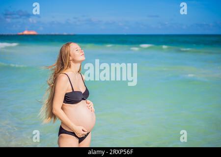 Radiosa donna incinta in costume da bagno, sullo sfondo mozzafiato di un mare turchese. Bellezza serena della maternità vicino alla riva Foto Stock