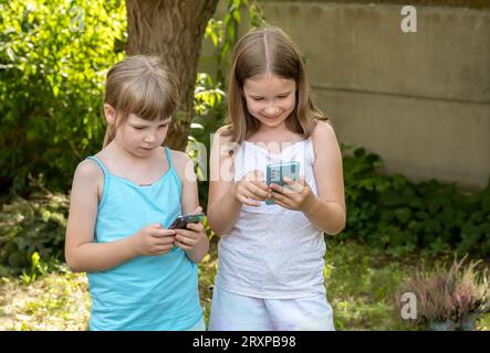 Ragazze bambini che tengono il loro smartphone in mano, bambini all'aperto che guardano i loro telefoni cellulari ridendo, usando tecnologia, app, scena esterna, reale Foto Stock