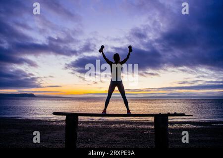 Silhouette di donna che si esercita con manubri sulla panchina costiera al crepuscolo nuvoloso Foto Stock