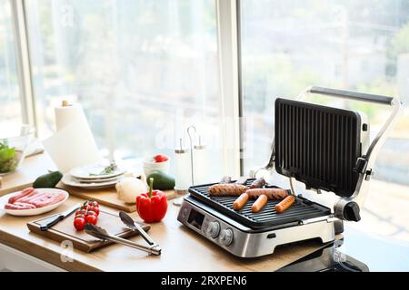 Moderna griglia elettrica con saporite salsicce su ripiano in legno in cucina Foto Stock