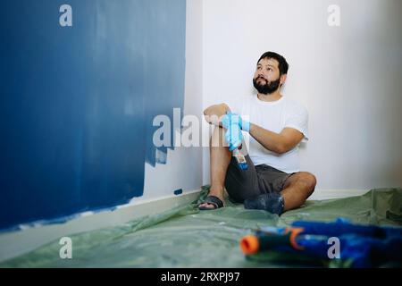 ristrutturazione della casa. Uomo barbuto caucasico che riposa dopo aver dipinto il muro immagine con messa a fuoco selettiva Foto Stock