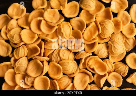 Pasta di orecchiette all'arancia essiccata sparsa su un tavolo di legno: Pezzi di orecchiette crude su fondo di legno scuro Foto Stock