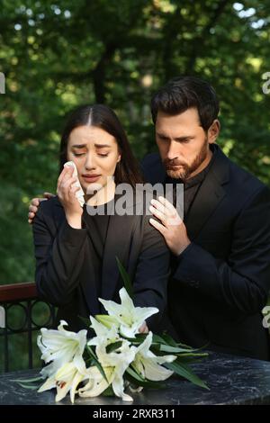Triste coppia in lutto vicino alla lapide di granito con gigli bianchi al cimitero. Cerimonia funebre Foto Stock
