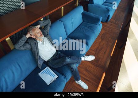 Un uomo d'affari anziano che si rilassa sul divano dopo una giornata di lavoro e guarda di lato Foto Stock