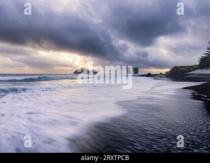 Lunga esposizione di onde sulla spiaggia di sabbia nera di Mosteiros con un tramonto nuvoloso e le pile di mare sullo sfondo, Mosteiros Foto Stock