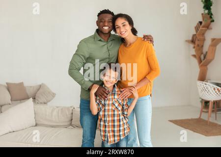 Genitori diversi che abbracciano il figlio sorridente a camera che posa a casa Foto Stock