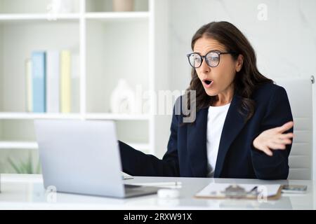 Stupita signora matura seduta in ufficio, guardando lo schermo del notebook Foto Stock