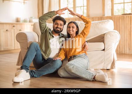 Felice giovane moglie e marito indiani seduti sul pavimento a fare il tetto con le mani, sorridendo alla macchina fotografica nell'interno di casa Foto Stock