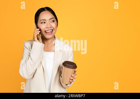 Giovane donna asiatica sorridente in completo con una tazza di caffè da asporto che parla sullo smartphone Foto Stock