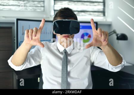 Trader Man con visore VR seduto in ufficio e guardando la tabella delle scorte virtuale. Foto Stock