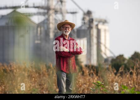 Ritratto di un agricoltore anziano con un cappello di paglia in piedi di fronte ai silos di grano nel campo di mais durante il raccolto Foto Stock