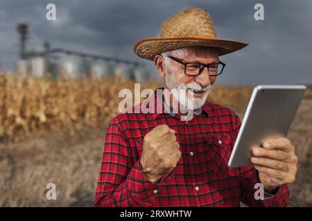 Un agricoltore entusiasta che guarda un tablet e celebra il successo sul campo davanti ai silos di grano durante il raccolto Foto Stock